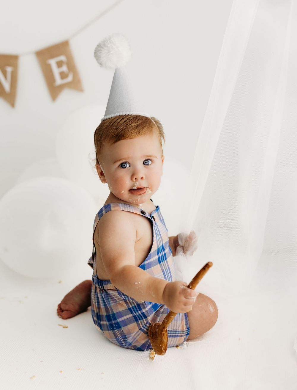 Adorable baby in plaid blue romper sitting on white backdrop with decorative bunting and holding wooden spoon. during a session in Fairfax,