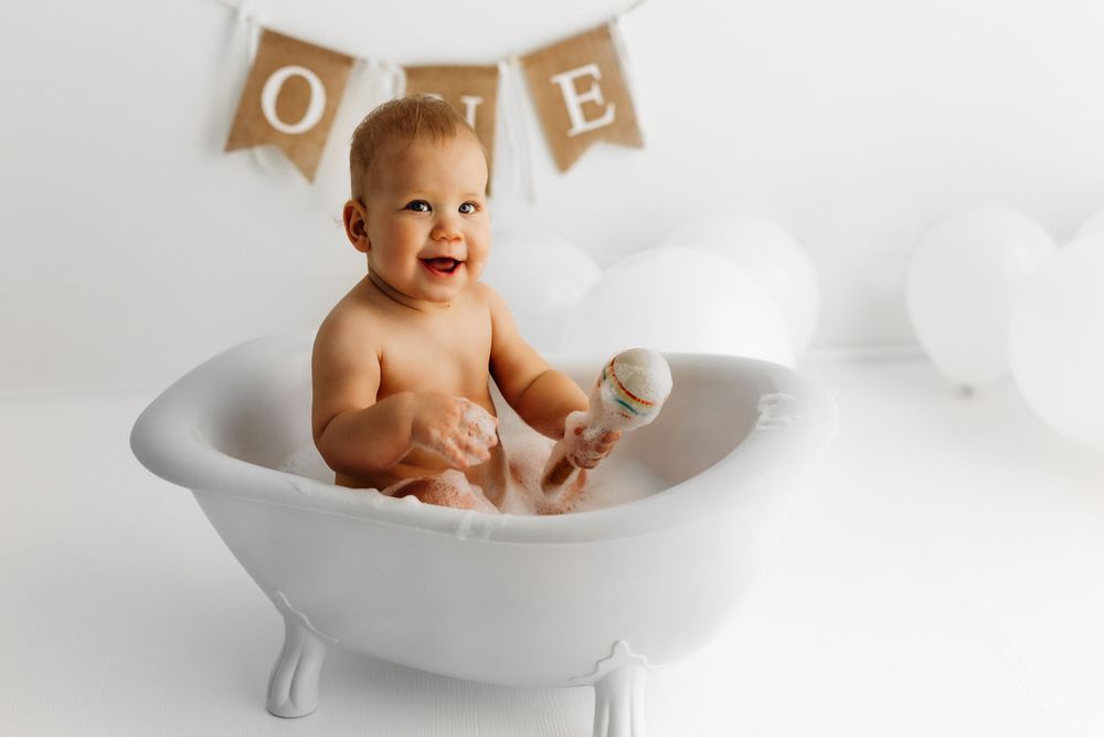 Happy baby sitting in miniature white bathtub with ONE banner decoration in background. during a session in Fairfax, VA.