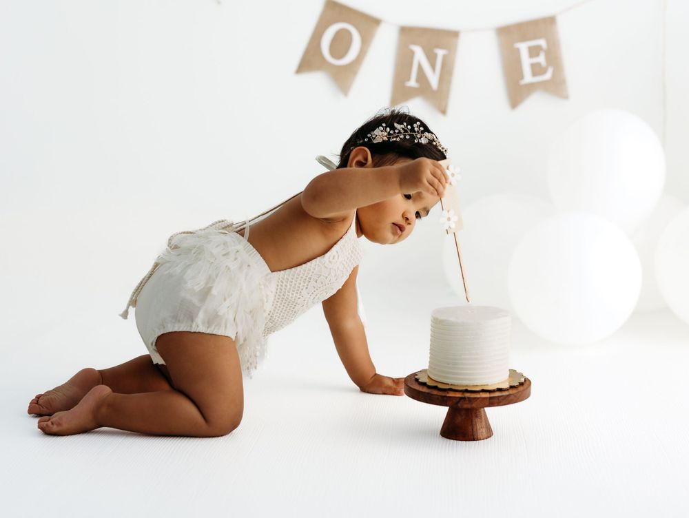 Baby in white outfit leans over to blow out candle on cake with ONE banner in background. during a session in Fairfax, VA.