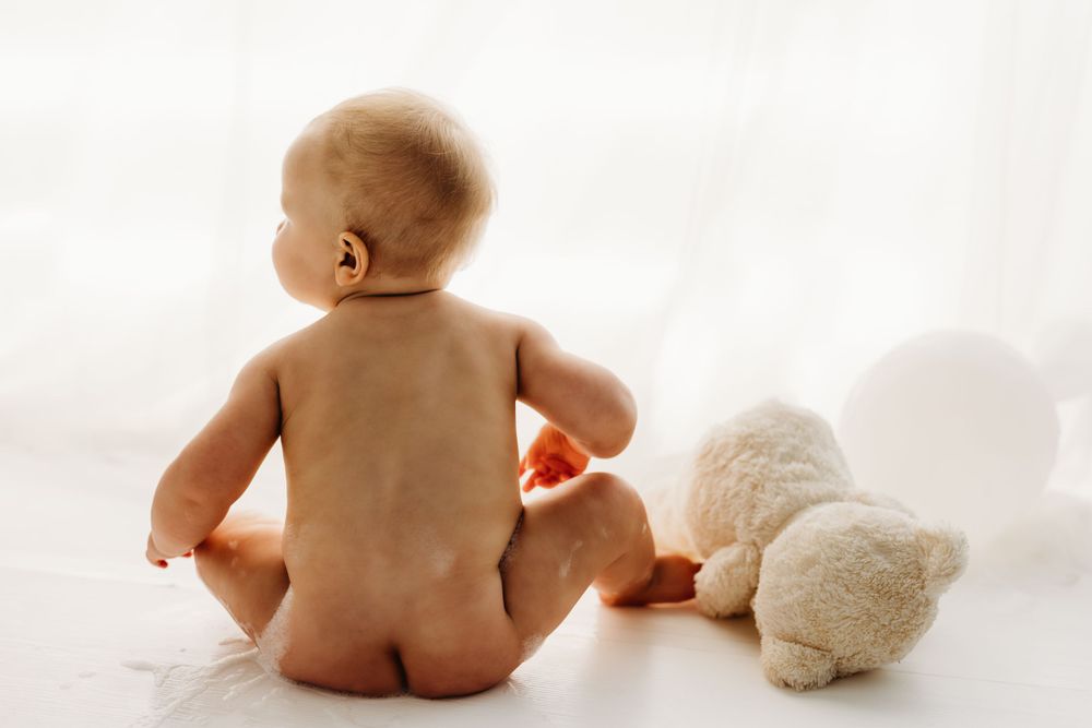 Back view of baby sitting on white floor next to plush teddy bear toy. during a session in Fairfax, VA.