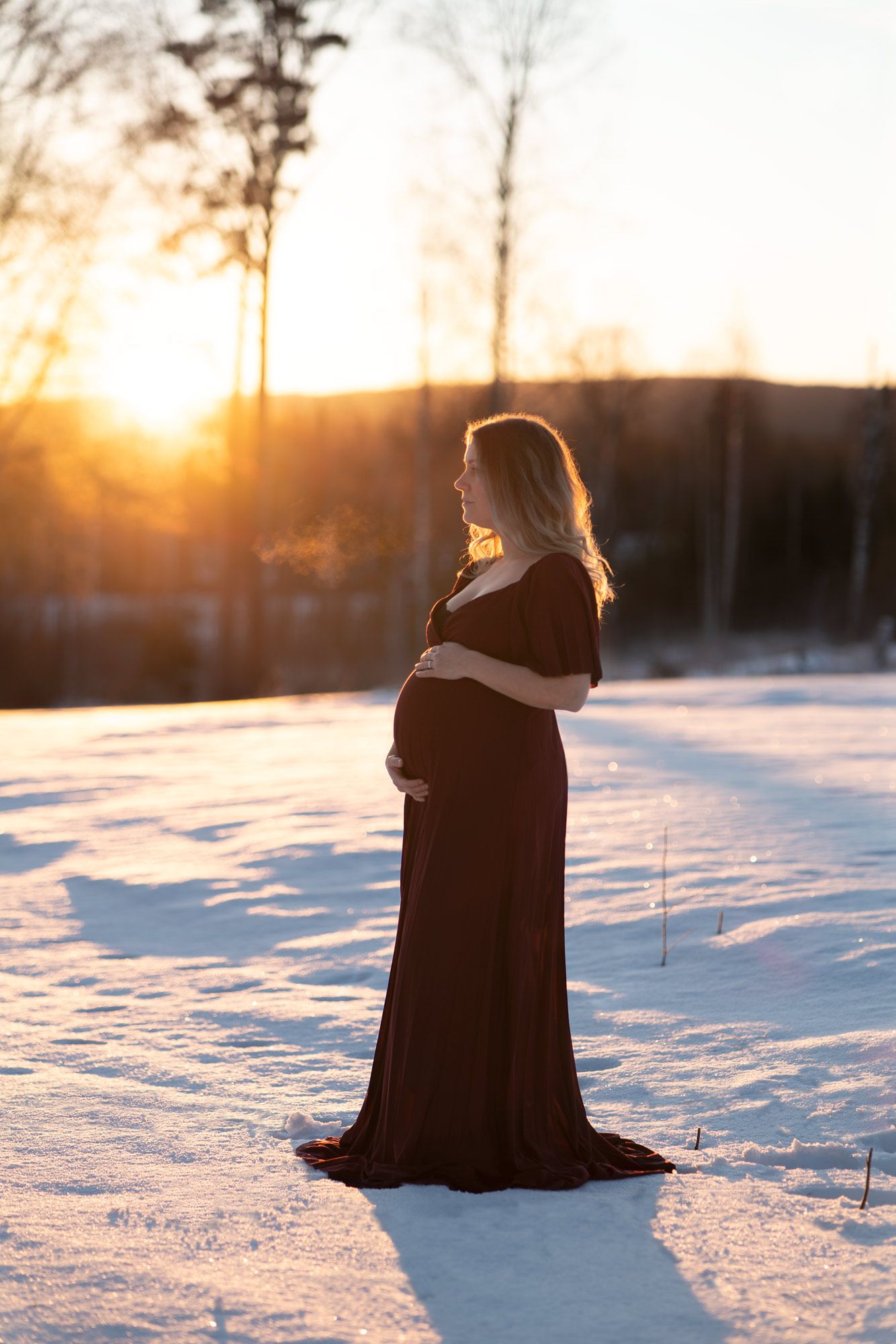 En gravid kvinna i vinröd långklänning håller om sin gravidmage i soluppgången. Det är en vintermorgon.