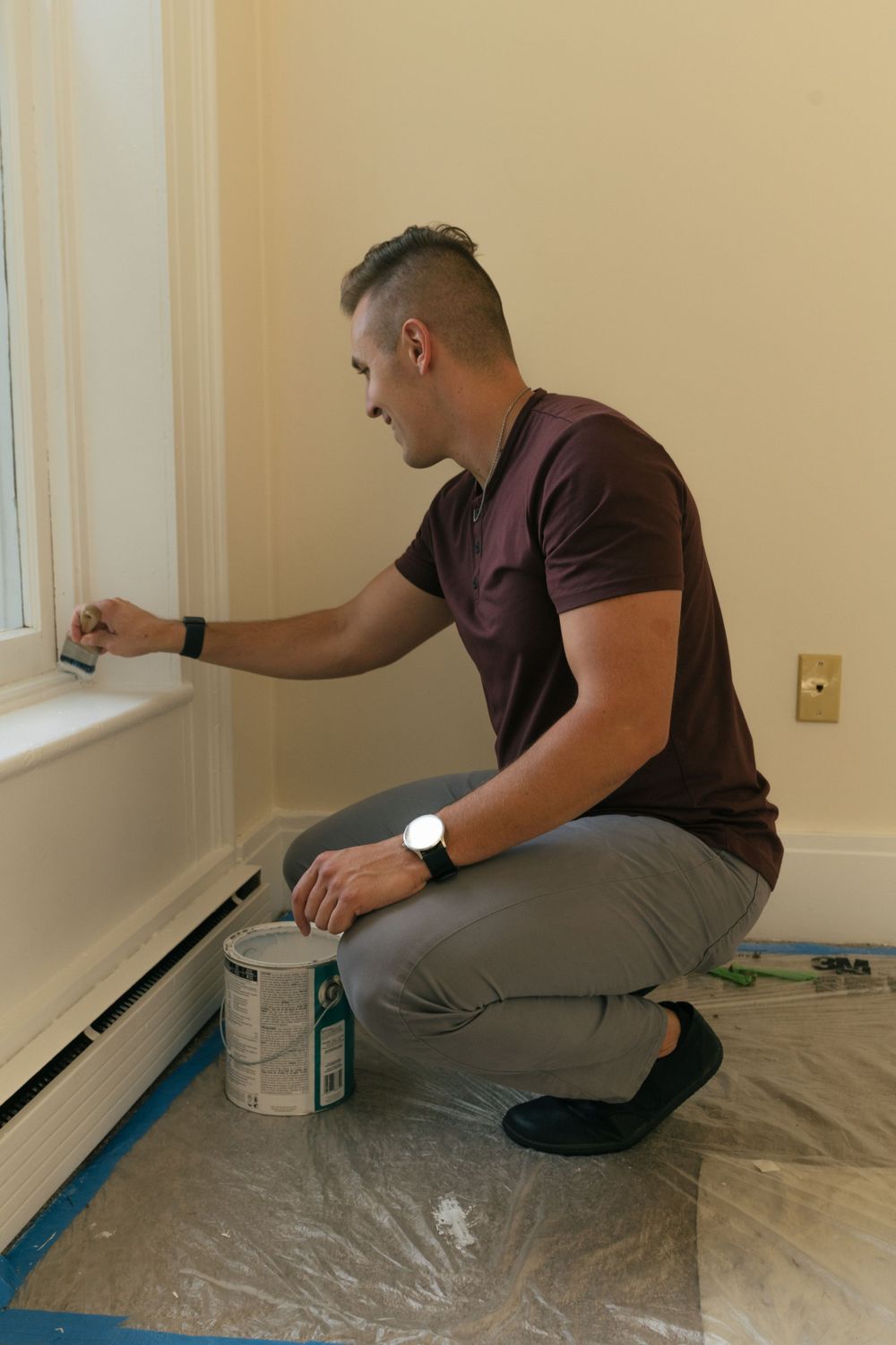 Person kneeling on protected floor painting baseboards in room under renovation.