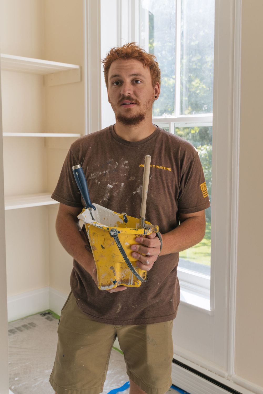 Worker in brown t-shirt holds yellow paint scraping tool while standing in room with white shelves.