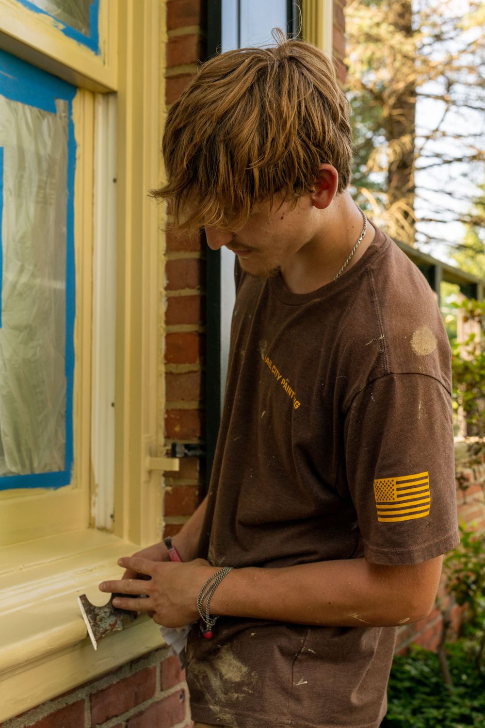 Person wearing brown t-shirt with flag patch working near a window.