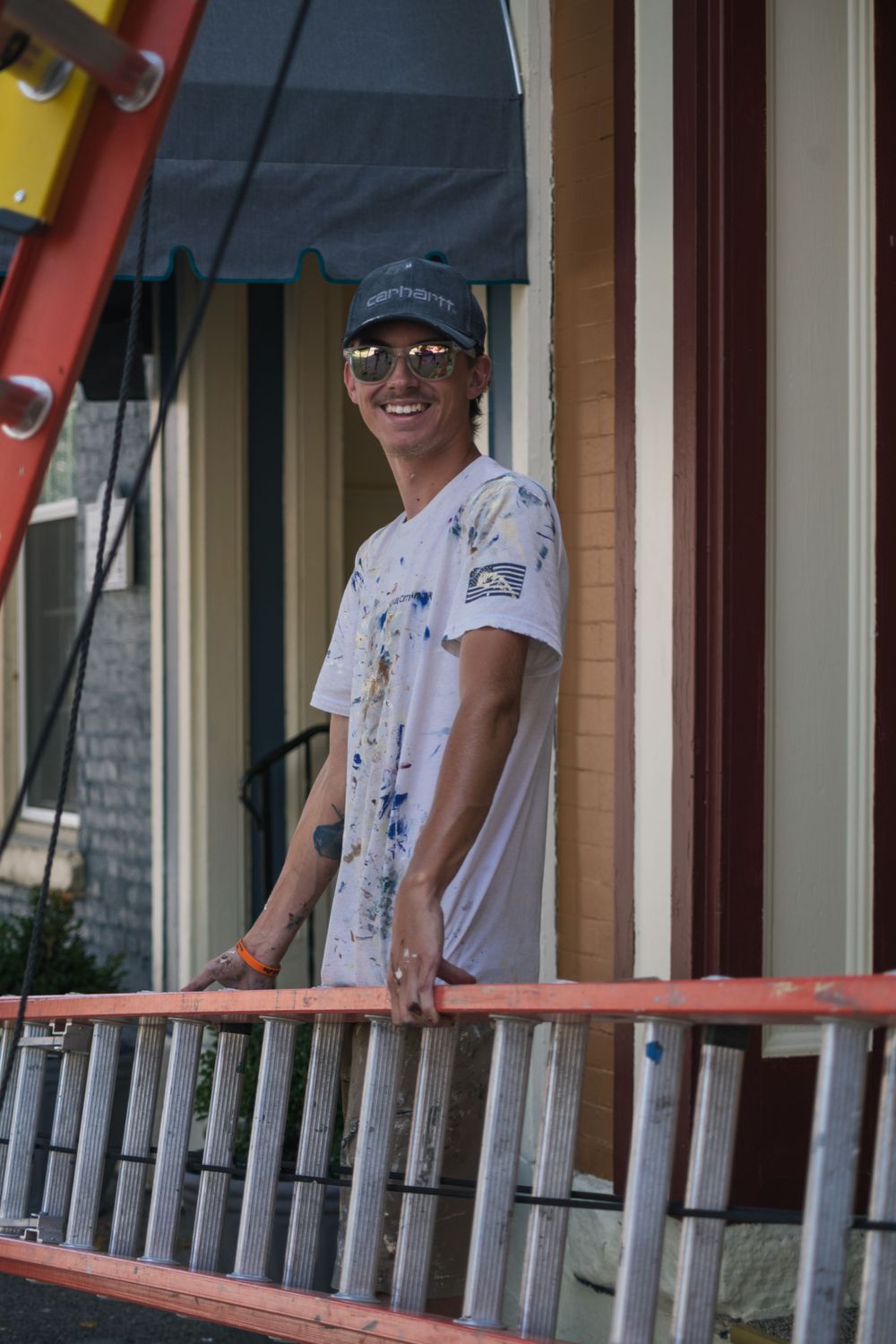 Person standing on porch near ladder wearing baseball cap and paint-splattered shirt.