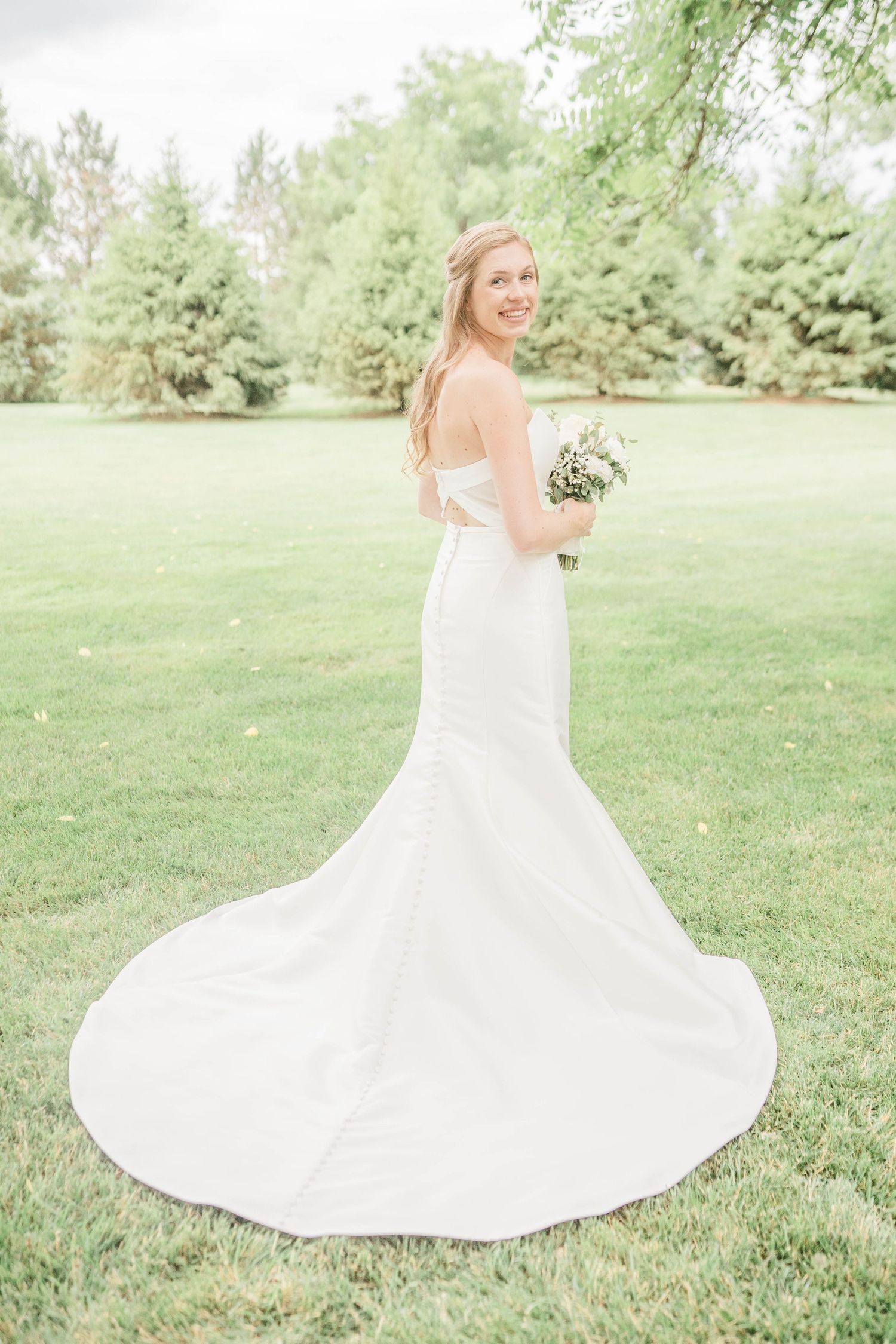 The image shows a bride standing alone in a grassy field, holding a bouquet of flowers.