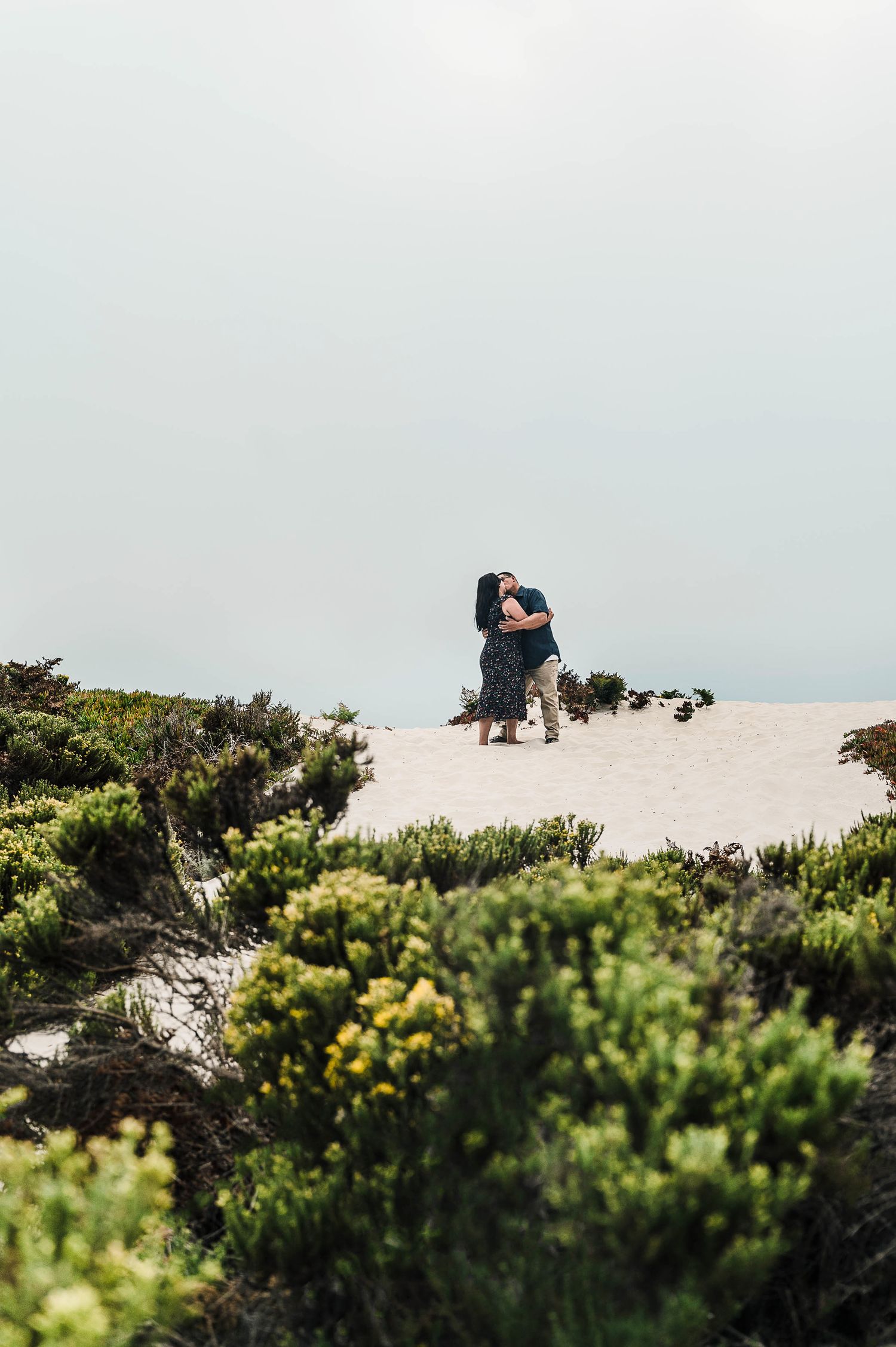 Couple embracing on a coastal sand dune surrounded by green shrubs.