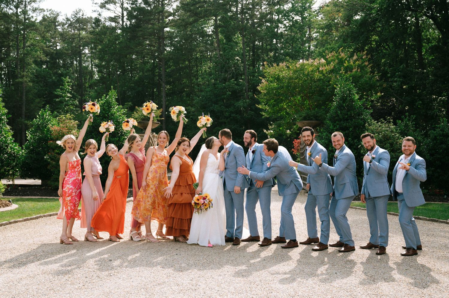 Wedding party in coordinated orange and blue attire poses together on a tree-lined driveway.