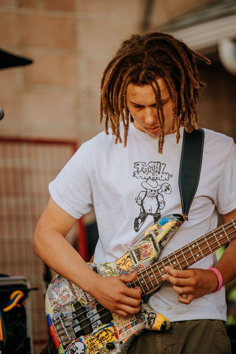 Musician with dreadlocks playing a sticker-covered bass guitar wearing a white graphic t-shirt.