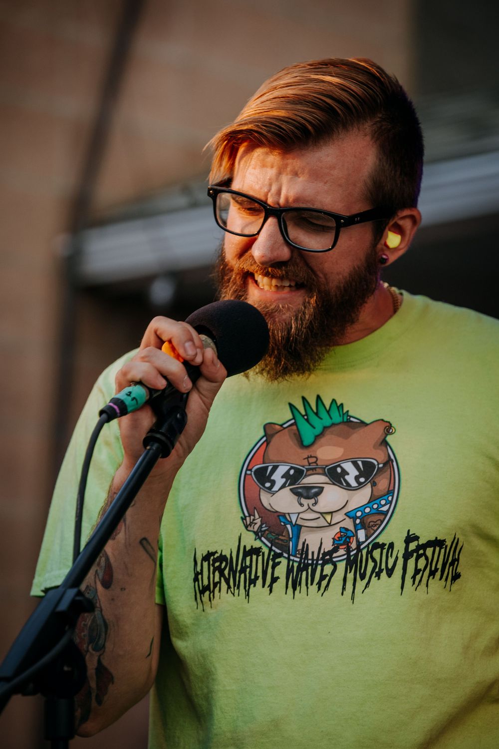 Singer with beard wearing neon green t-shirt performing on outdoor stage.