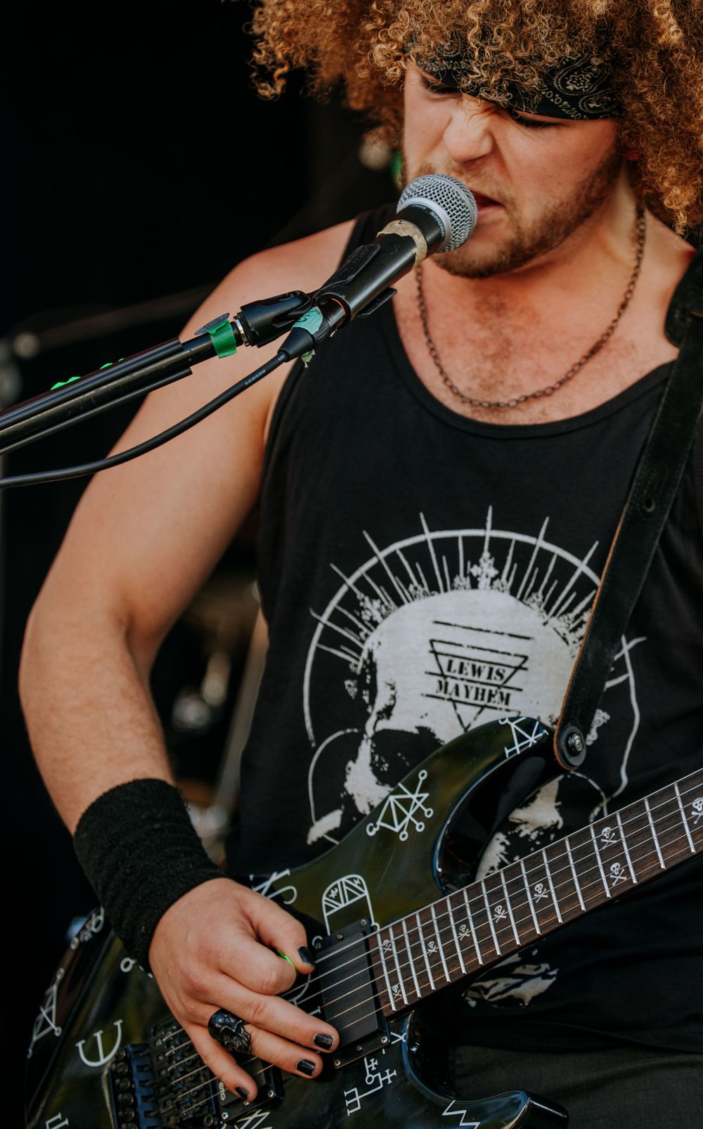 Musician with curly hair playing guitar and singing into microphone wearing black tank top.
