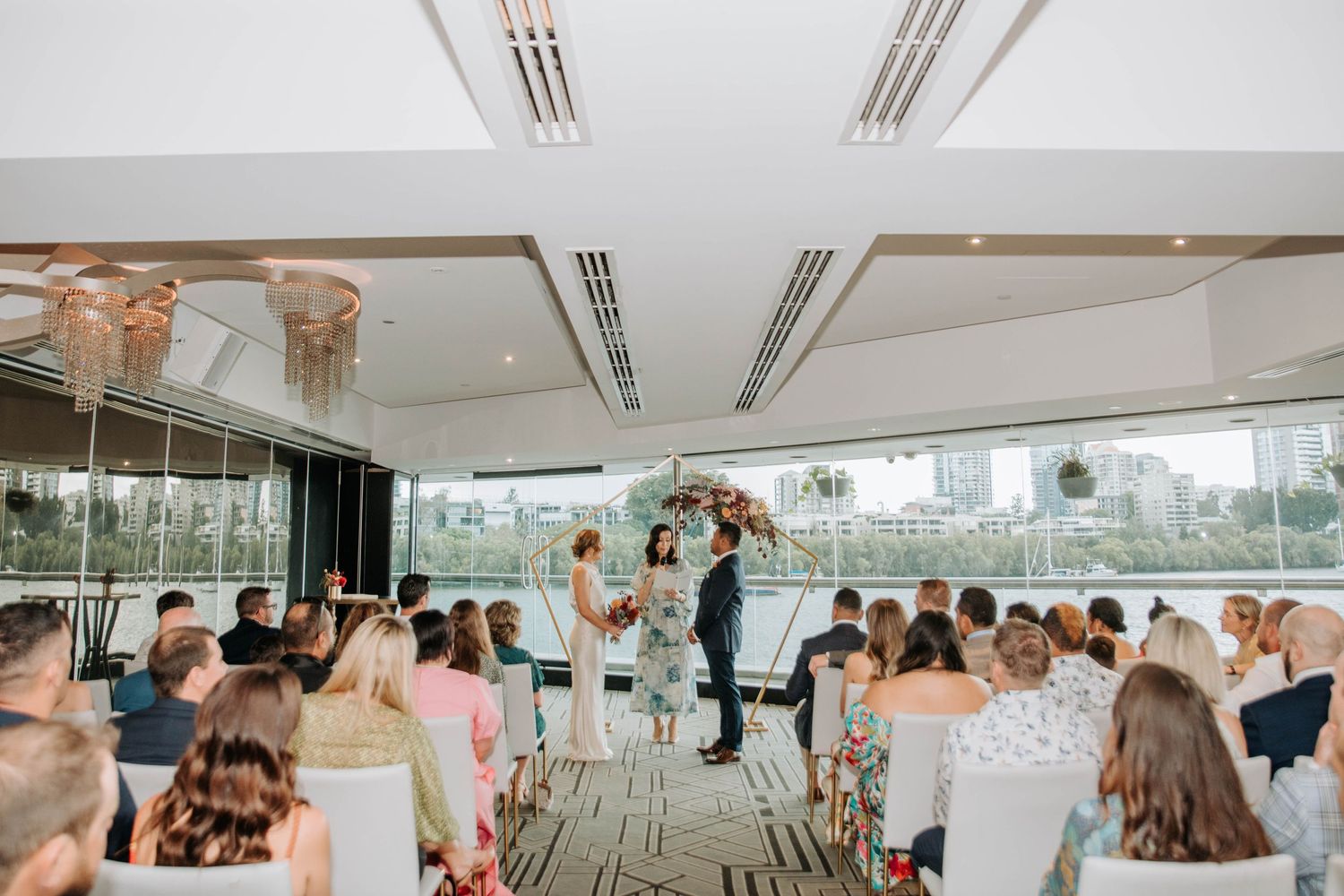 A wedding ceremony in progress in a modern glass-walled venue with guests watching.
