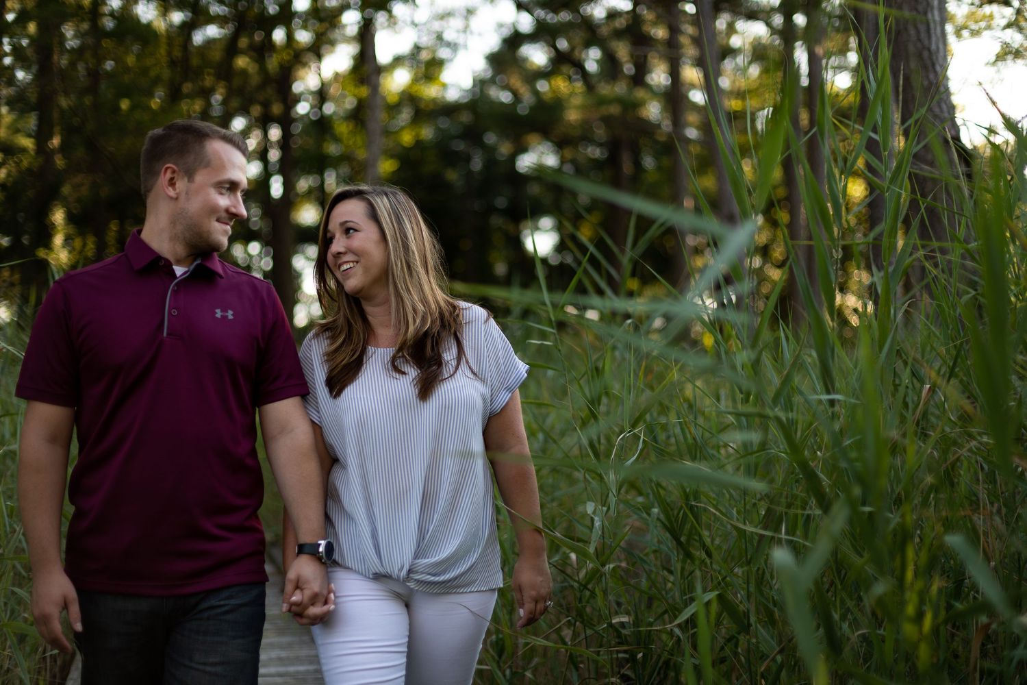 Couple holding hands while walking along wooden path through tall grasses.