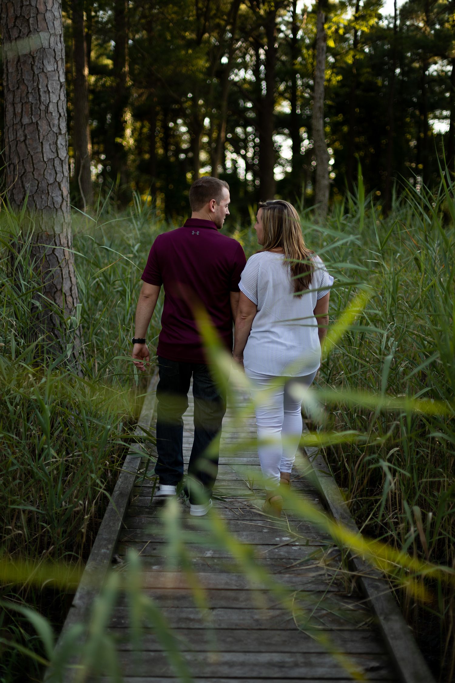 Couple walking together on wooden boardwalk through tall grass and trees at dusk.