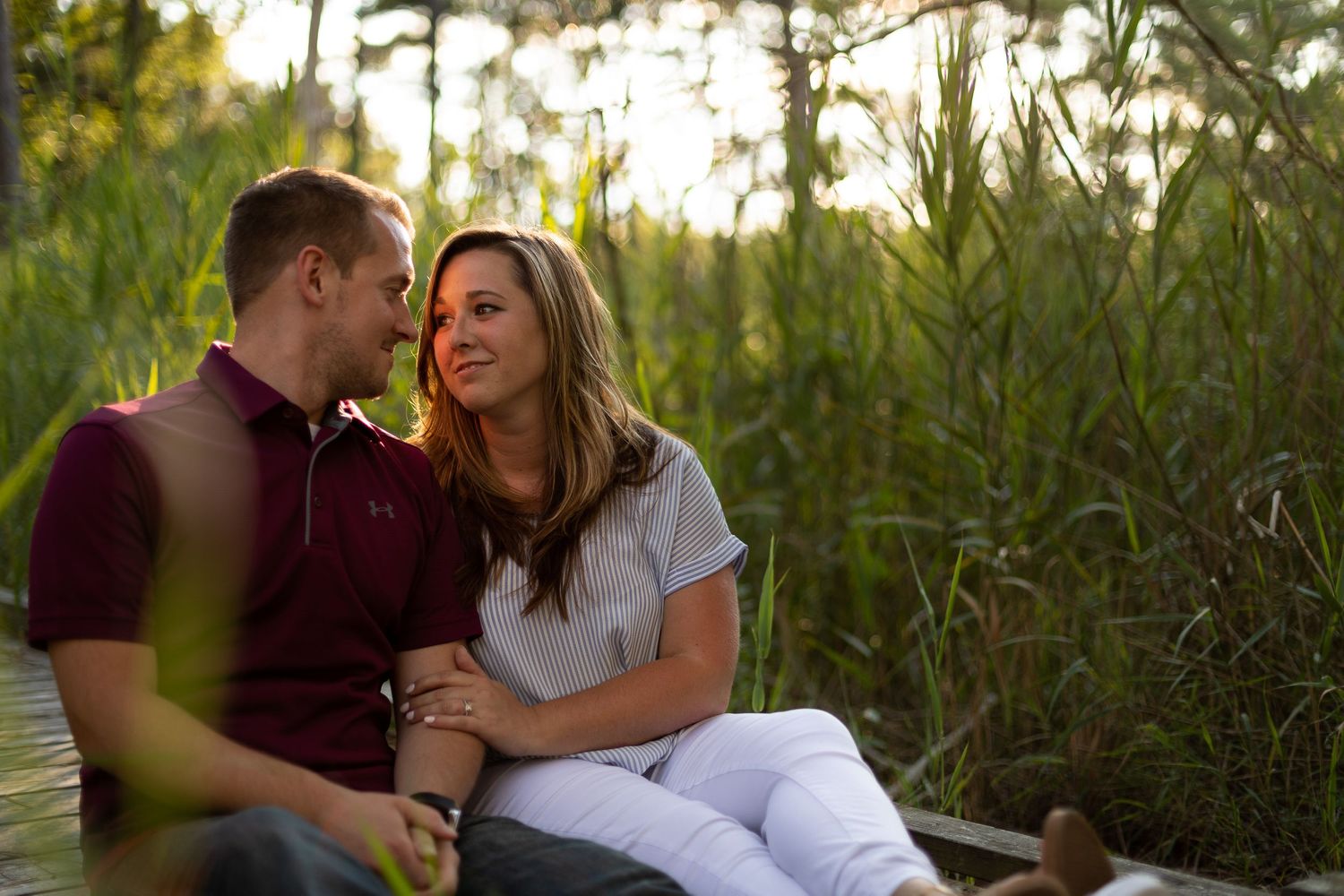 Romantic couple shares intimate moment on wooden boardwalk surrounded by tall grass at sunset.