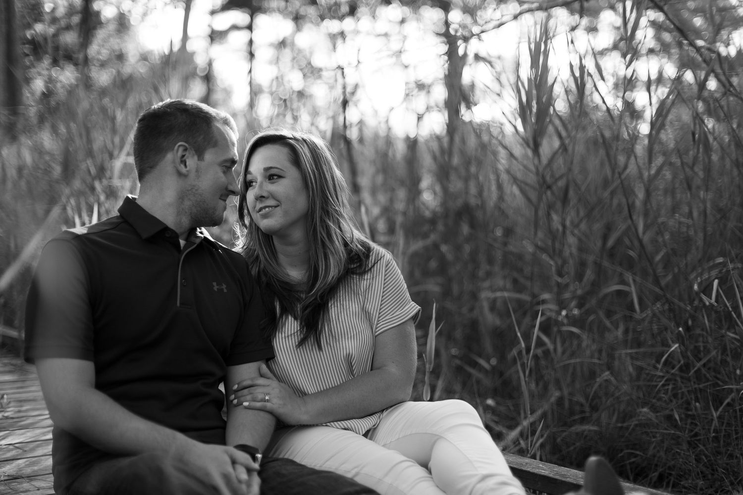 Black and white portrait of couple sharing tender moment on wooden walkway among tall grasses.