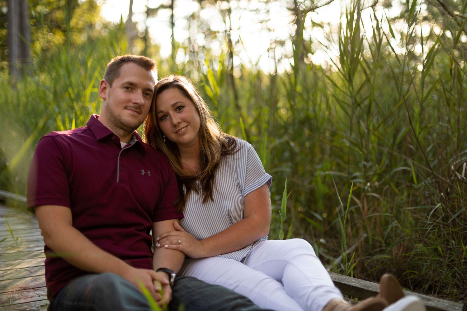 Couple in casual attire sitting close together on wooden path among marsh grass at golden hour.