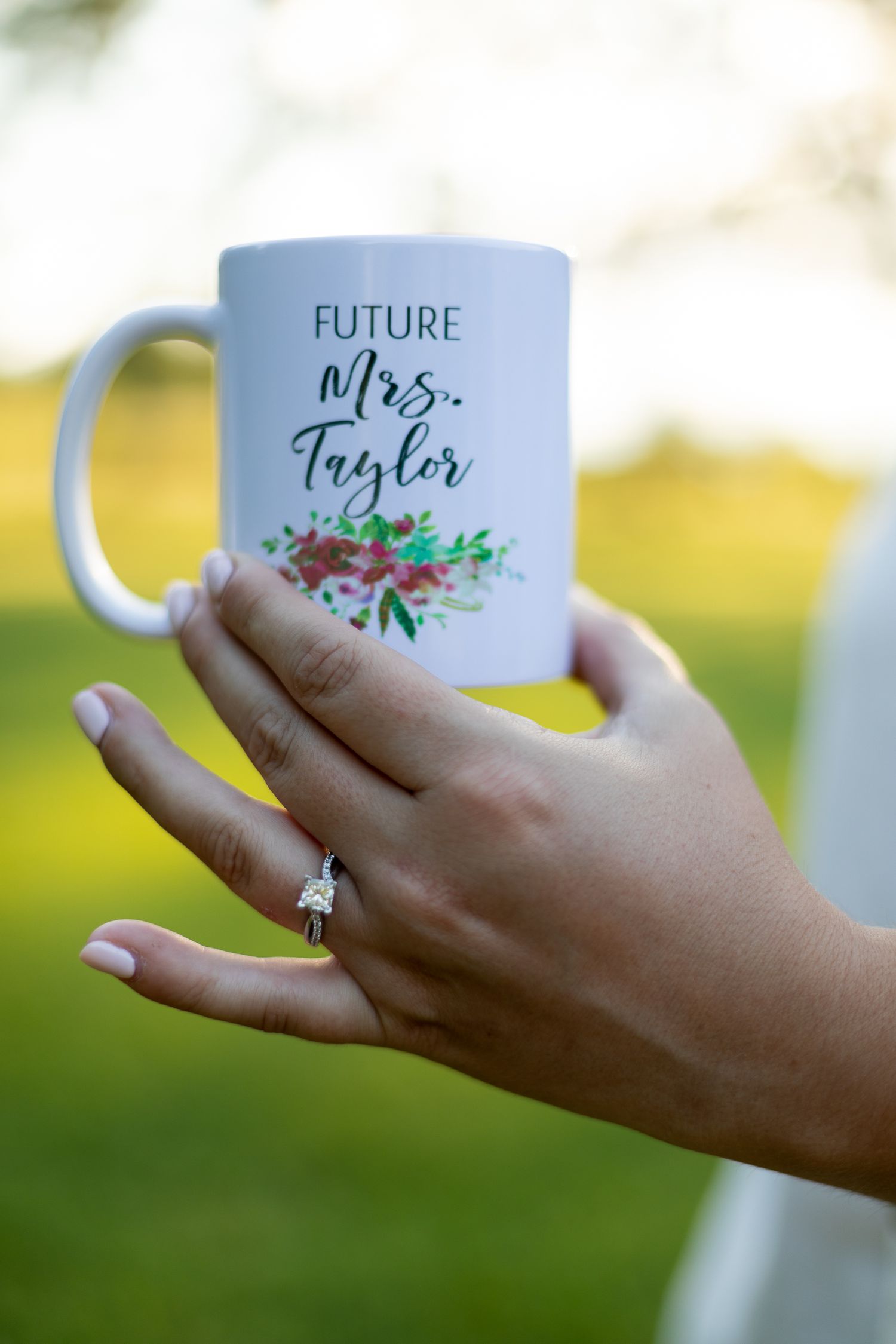 White coffee mug with 'Future Mrs. Taylor' text and floral design held by hand with engagement ring.
