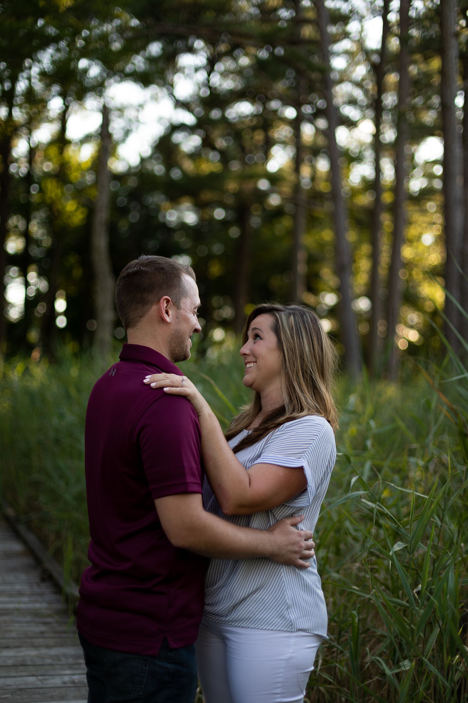 Couple embraces on wooden boardwalk surrounded by tall grass during sunset.