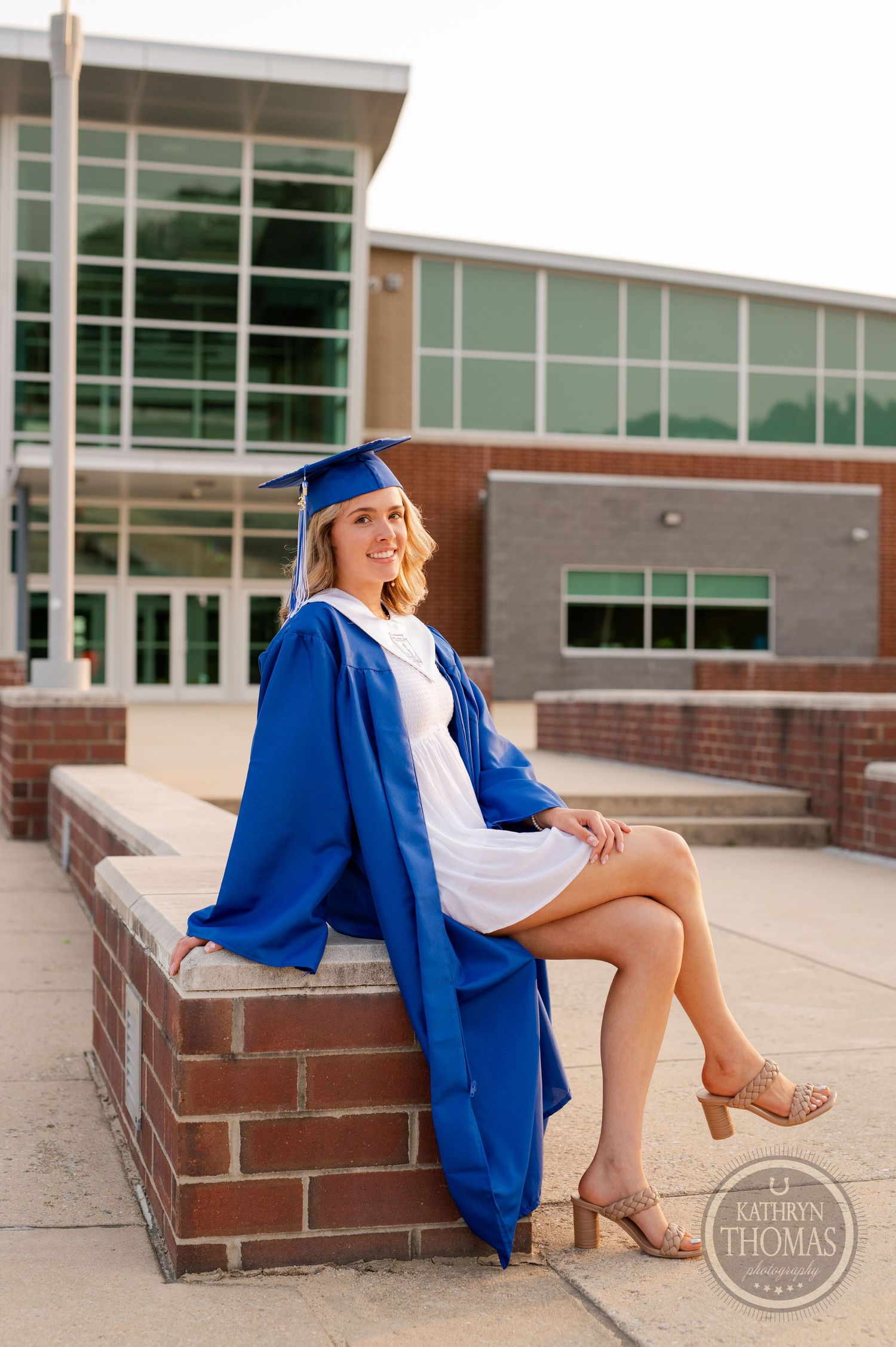 Graduate seated on brick wall wearing blue graduation gown and white dress.