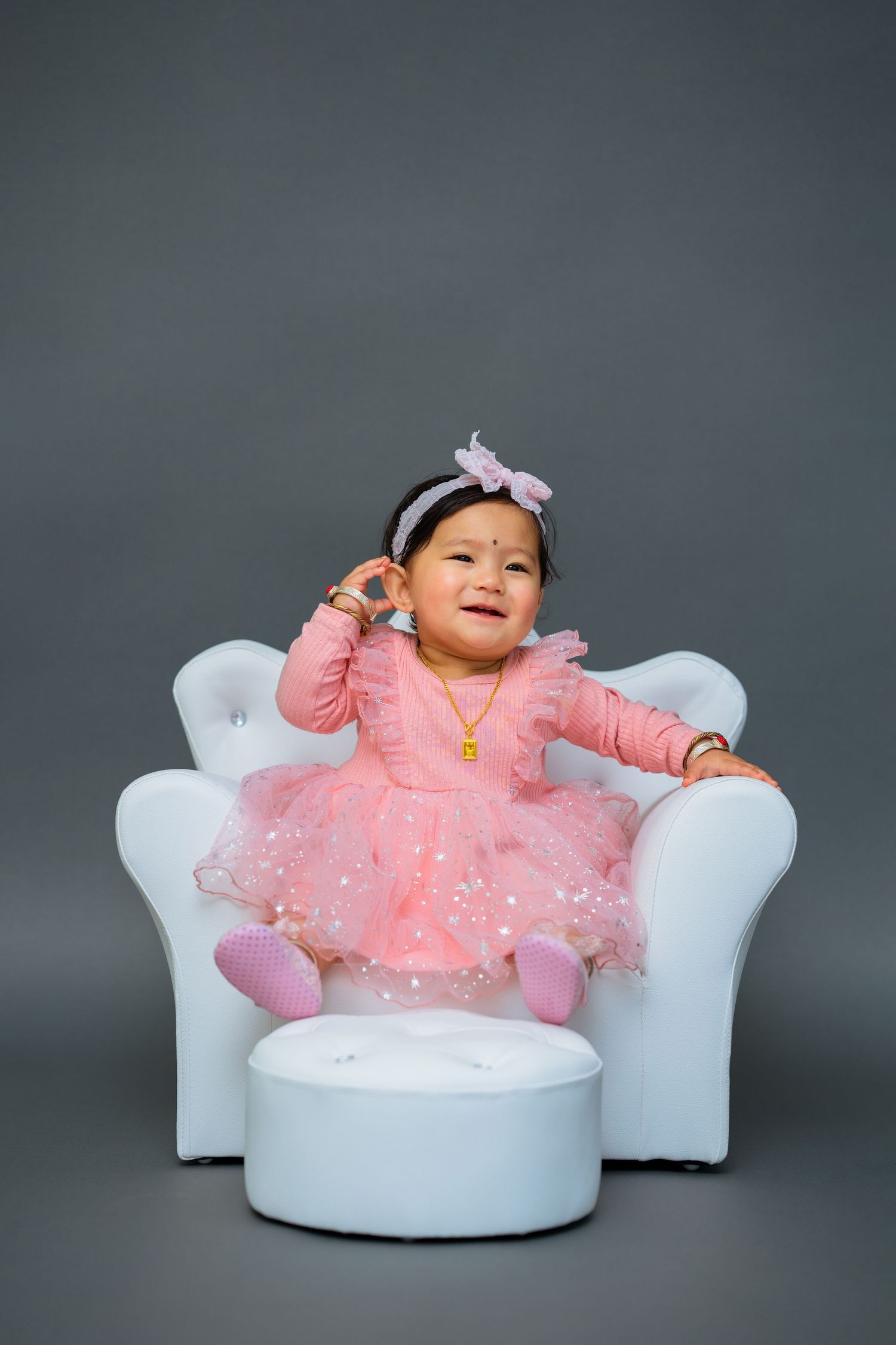 Happy baby in pink tutu dress sitting on white chair against grey background.