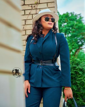Sophisticated navy pinstripe suit with belted waist, white hat and sunglasses against brick wall.