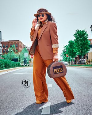 Stylish brown pantsuit with designer bag and patterned visor on city street.