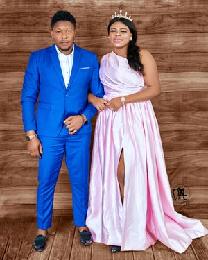 Couple in formal attire poses together against wooden backdrop at celebratory event.