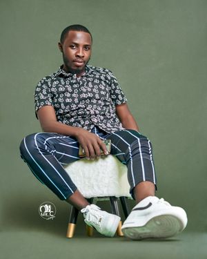 Person in floral shirt and striped pants sits casually on white stool against olive green background.