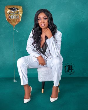 Person in a white pantsuit sits elegantly on a stool with a gold graduation balloon against a teal backdrop.