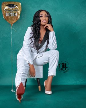 Stylish graduate poses in white suit against teal background with metallic balloon decoration.