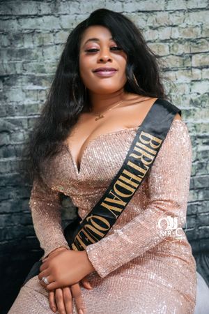 Woman wearing sparkly rose gold dress and birthday sash poses against brick wall.
