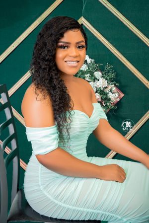 Woman in mint green dress poses elegantly on chair with floral decoration behind.