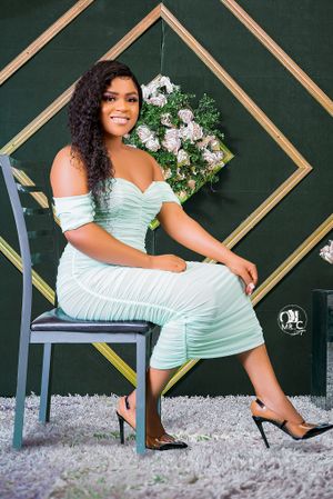 Woman in mint green ruched dress poses on chair against geometric backdrop with floral decorations.