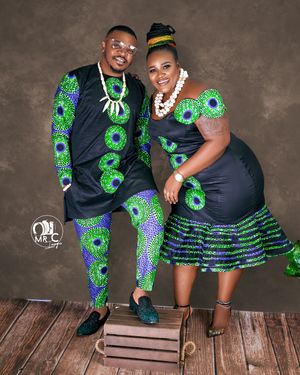 A couple wearing coordinated navy and green African print outfits pose together on wooden platform.