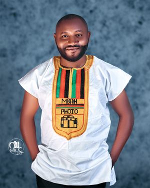 A professional photographer wearing a white shirt with decorative African-style collar patch stands against blue backdrop.