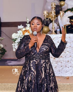 A performer in an elegant black lace gown sings into a microphone at an event with floral decorations.