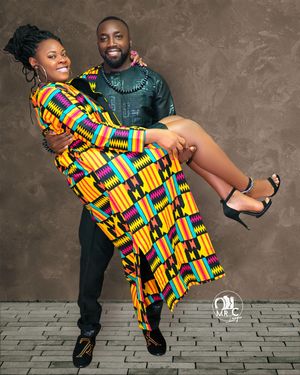 A couple in traditional African attire poses playfully with bright kente cloth patterns against a textured brown wall.