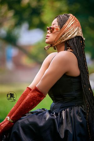Artistic portrait in a black dress and red arm warmers with braided hair against a blurred outdoor background.