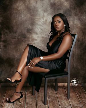Elegant portrait of woman in black dress seated on chair with wooden floor background.