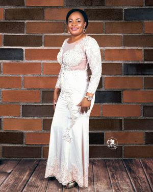 Elegant full-length portrait in white lace gown against brick wall background.