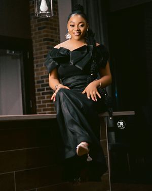 Woman in elegant black off-shoulder dress poses against brick wall with moody lighting.