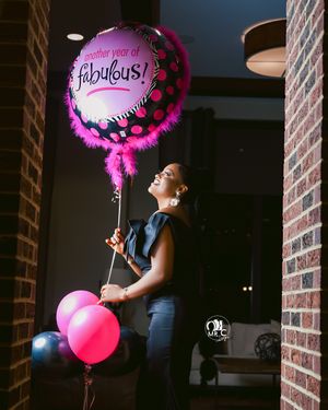 Side view of person holding pink 'fabulous' balloon and other party balloons in brick hallway.