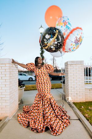 Joyful celebration pose in orange patterned mermaid dress holding festive balloons between brick pillars.