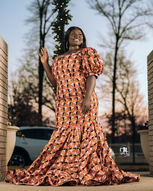 Stunning orange patterned mermaid gown with puff sleeves photographed at sunset with a Christmas tree in background.