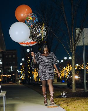 Person in geometric print dress holds festive balloons outdoors at night with lit trees in background.