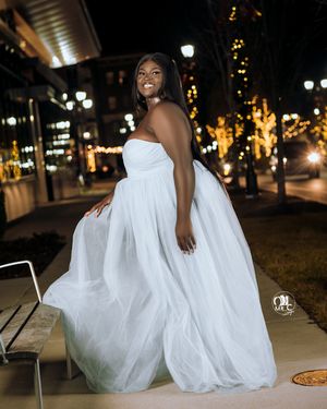 Woman in flowing white gown poses elegantly on city street at night with illuminated trees.