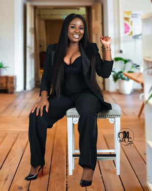 Professional wearing all black business attire poses on white stool in bright hallway.
