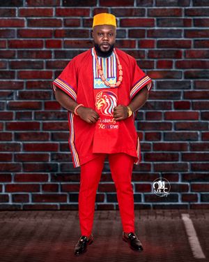 A person in red traditional Nigerian attire with yellow cap poses against a brick wall.