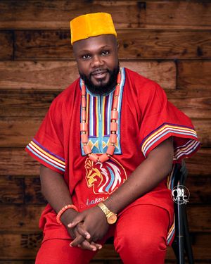 A person wearing a red traditional outfit and yellow cap sits against a wooden wall background.